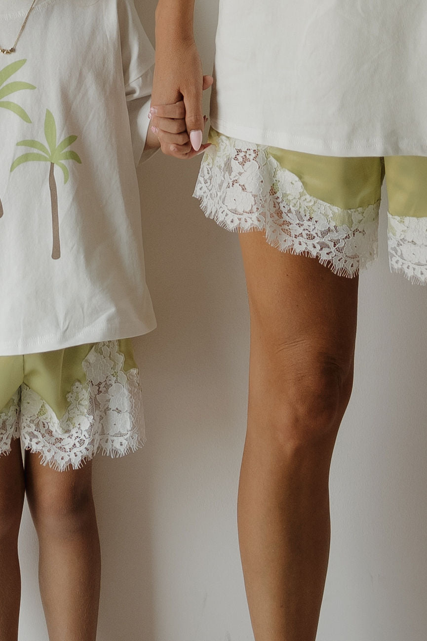 An adult and a child hold hands, both wearing relaxed fit matching “Women’s Satin Shorts | Prickly Pear” in light green with white lace trim from forever french baby. The child wears a palm tree shirt, while the adult wears a plain white shirt. Only their lower bodies are visible.