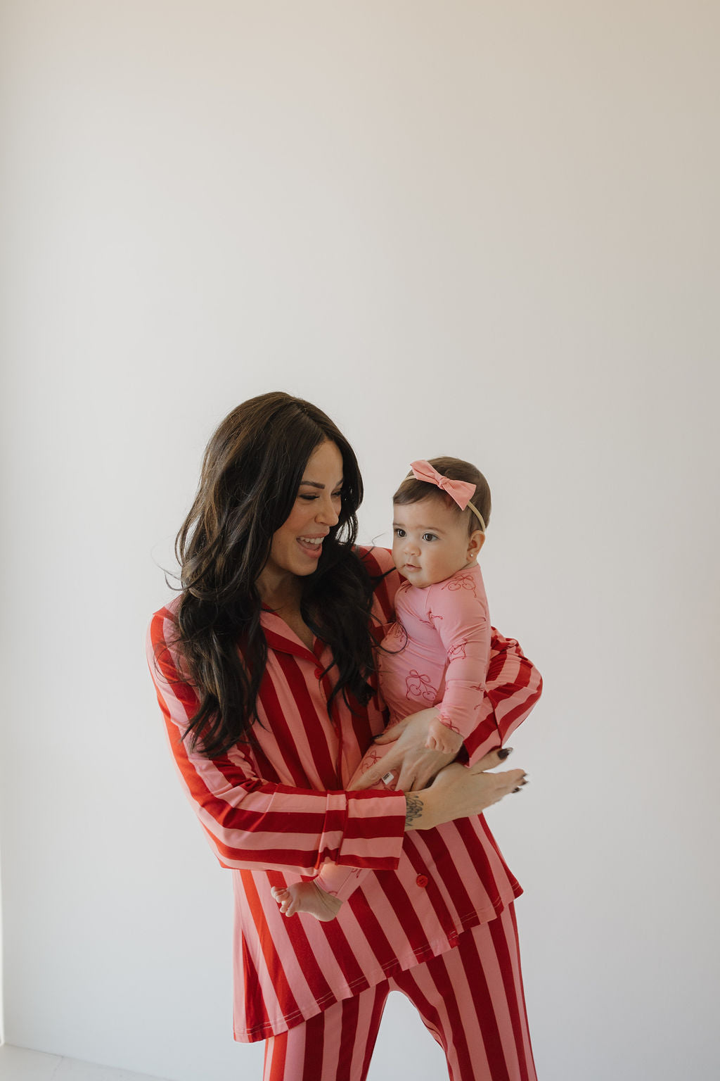 A woman wearing the Minty Cherries Women's Bamboo Button Up Lounge Set in Iconic Stripe smiles at a baby in pink with a matching headband, both standing before a plain light background.