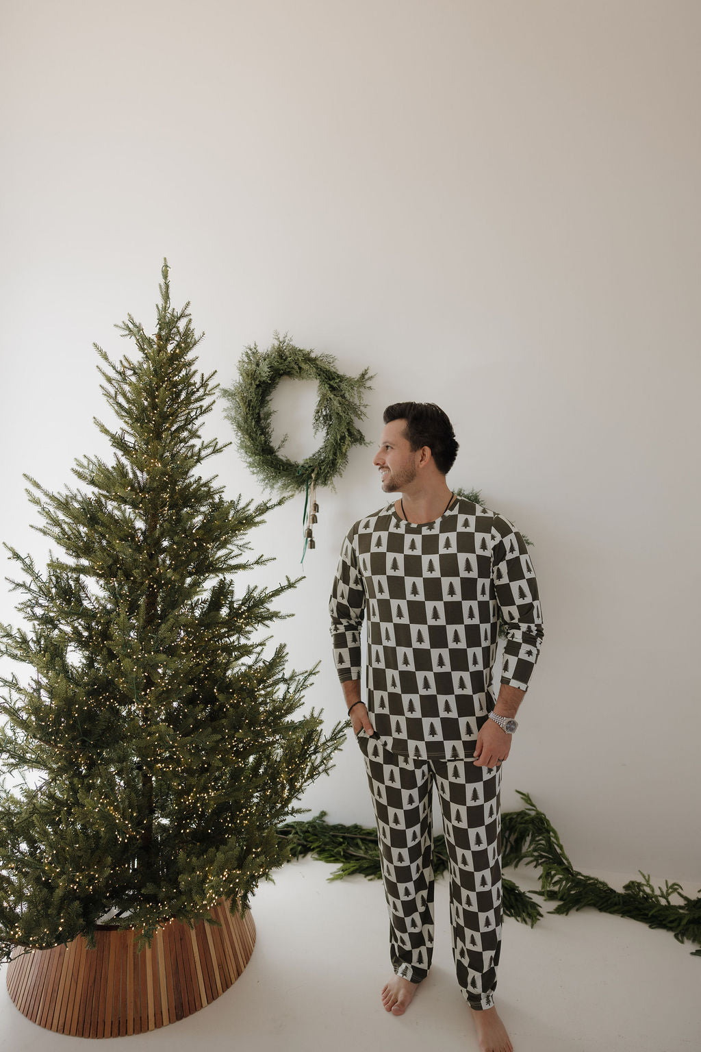 A man in forever french baby Men’s Bamboo Pajamas | Christmas Tree Checkerboard stands barefoot by a decorated Christmas tree, gazing at a wreath on a plain white wall.