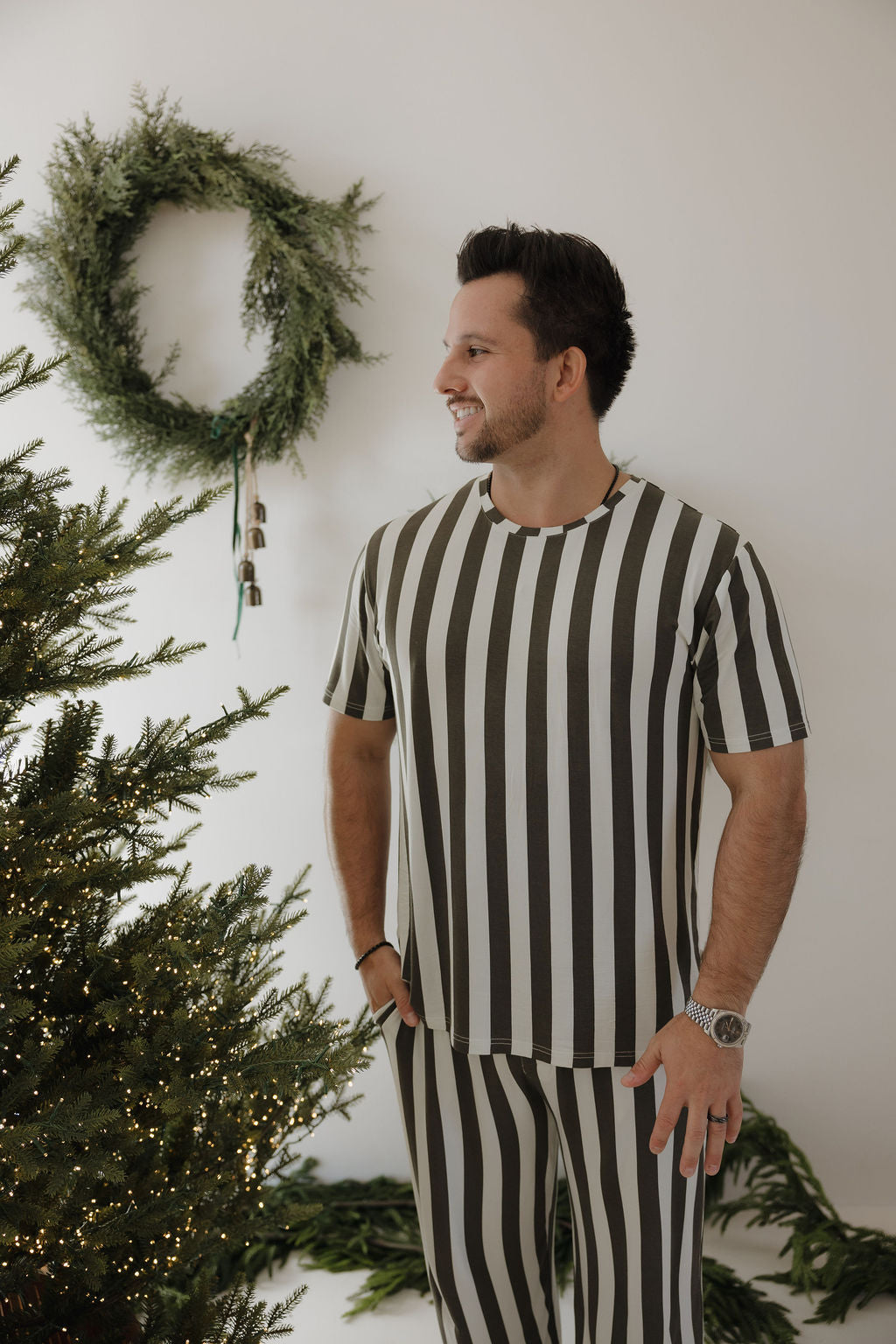 A man in forever french baby's Men's Short Sleeve Bamboo Pajamas in Fireside Stripe stands by a Christmas tree, smiling, with a green wreath on the white wall behind him.