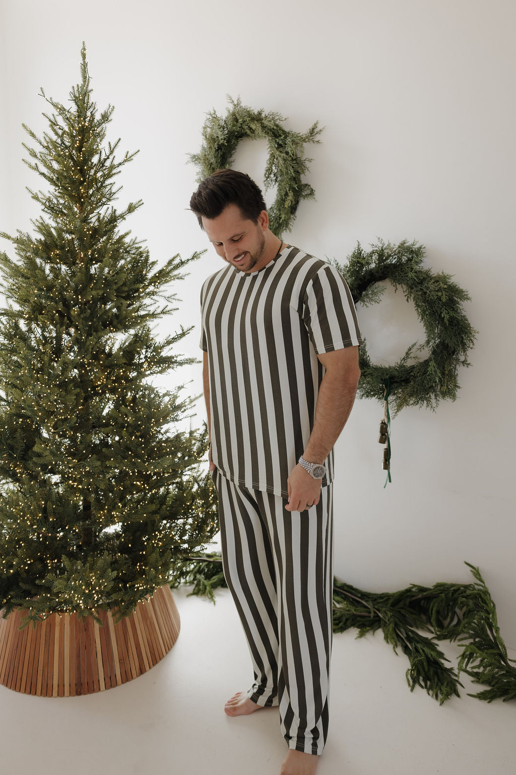 A man wearing forever french baby's Men's Short Sleeve Bamboo Pajamas in Fireside Stripe stands barefoot, smiling by a decorated Christmas tree, with two green wreaths hanging on a white wall behind him.