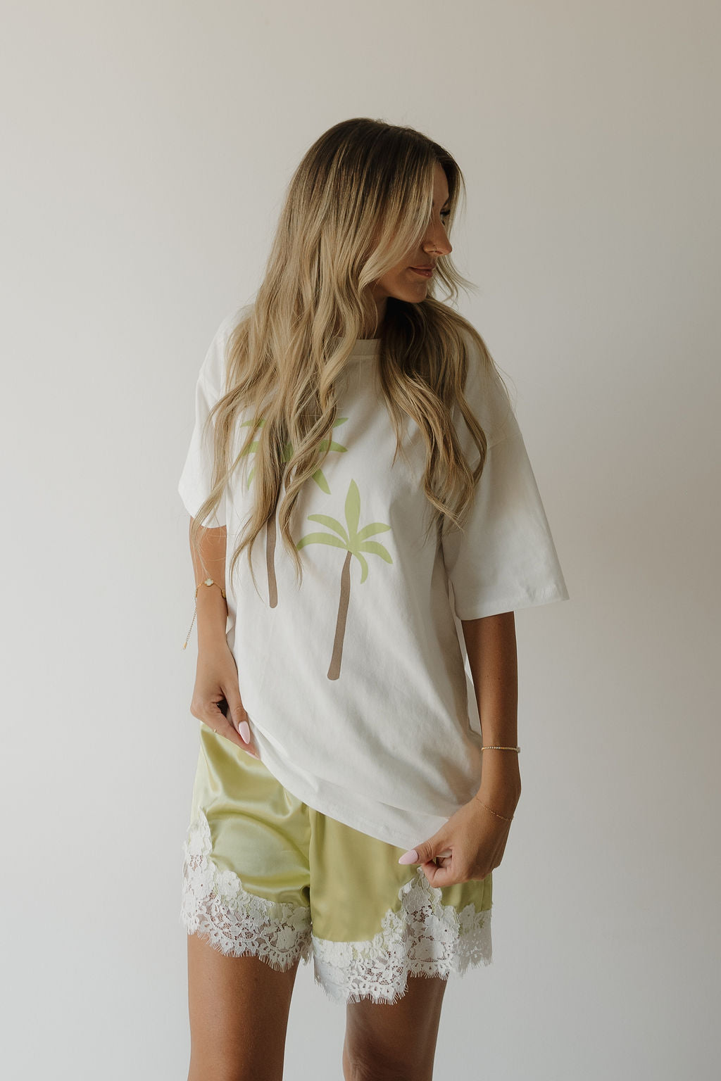 A woman with long blonde hair wears the forever french baby Adult Cotton Tee | Palm, paired with light green satin shorts trimmed with white lace, standing against a plain background.