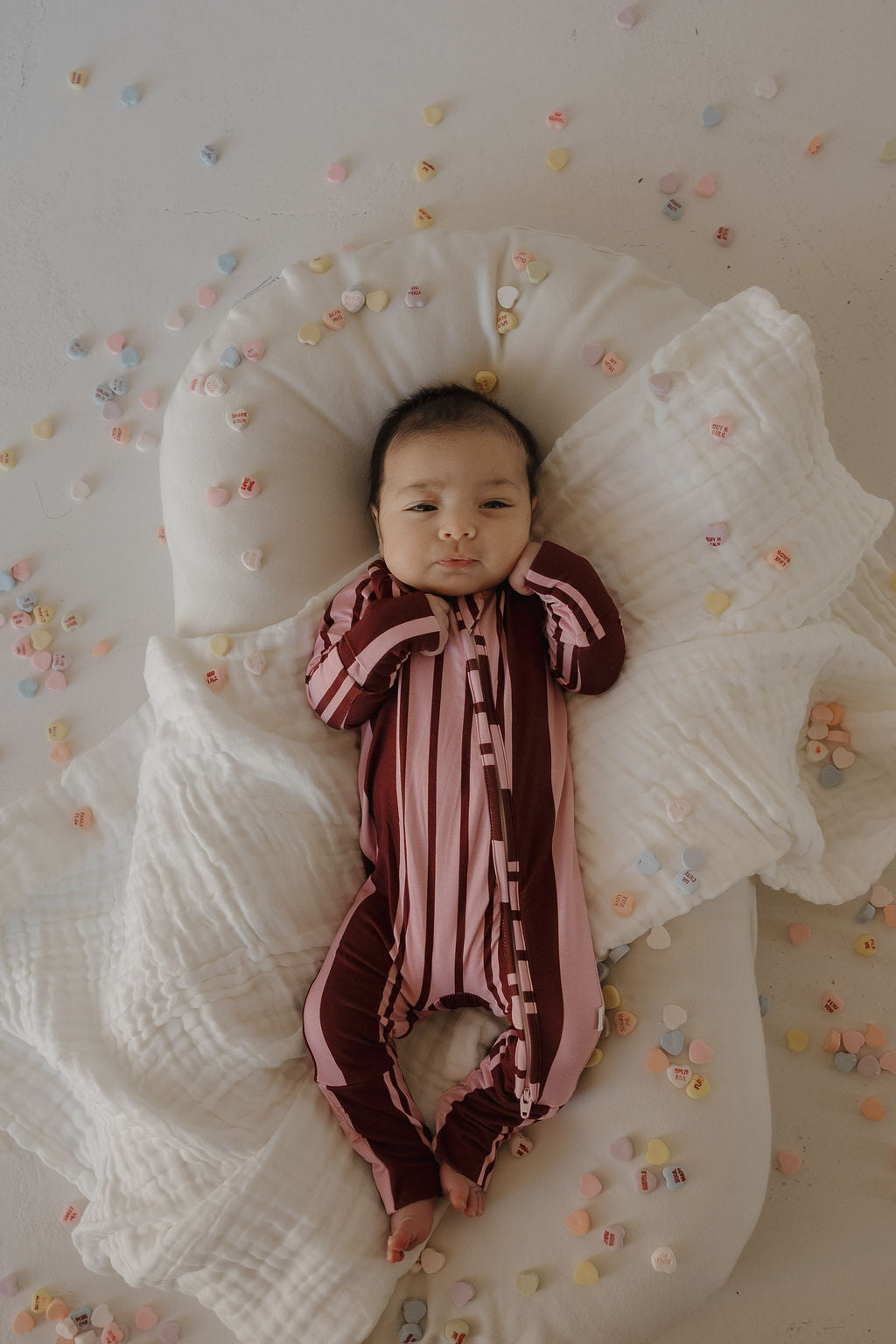 A baby wearing forever french baby's Bamboo Zip Pajamas in Sweetheart Stripes rests on a white cushion with a quilted blanket, surrounded by colorful candy hearts scattered on the floor.