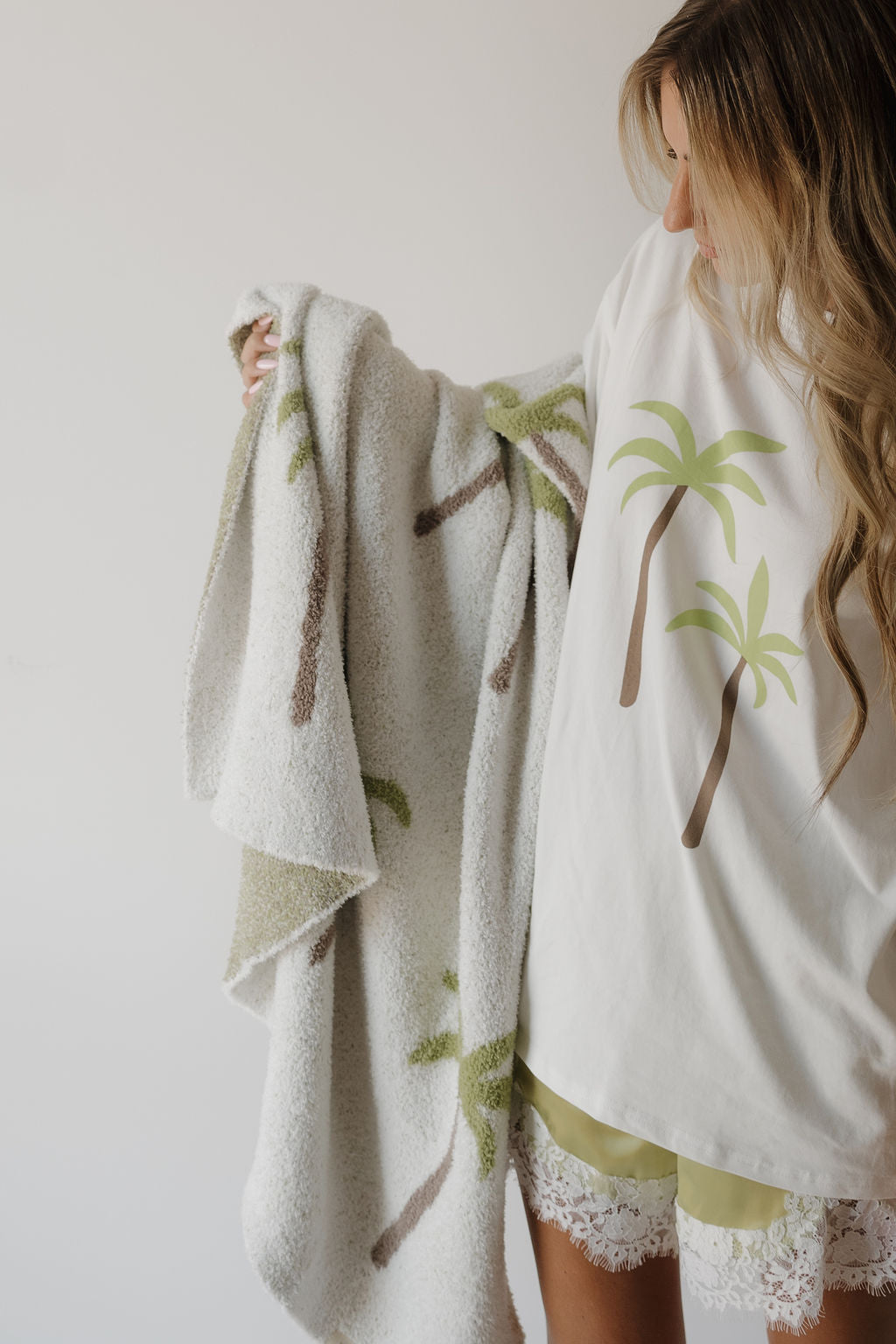 Wearing a palm tree T-shirt and lace shorts, a woman holds the forever french baby Plush Blanket | Palm—a soft, Oeko-Tex certified blanket featuring a matching palm tree print—while standing against a plain light background.