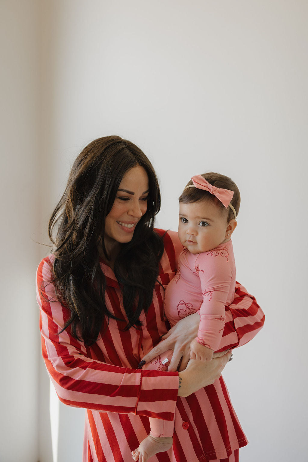 A woman in a red and pink striped dress holds a baby wearing Minty Cherries' Bamboo Zip Pajamas | Iconic Cherry, paired with a matching pink bow headband. They stand against a light background as the woman smiles at the baby.