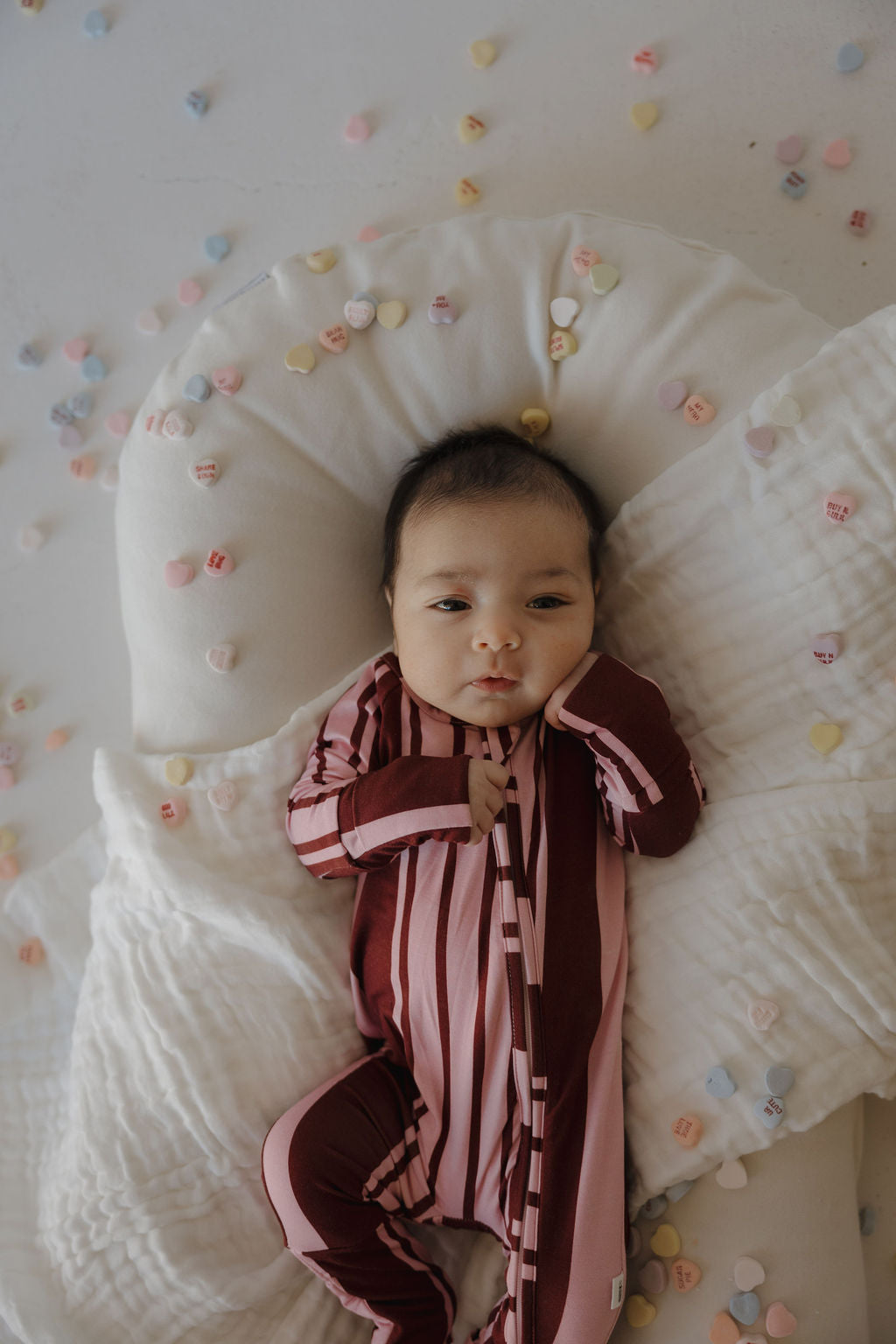 A baby in forever french baby Bamboo Zip Pajamas | Sweetheart Stripes lies on a white blanket, surrounded by colorful heart-shaped candies, gazing up with one hand near their cheek.