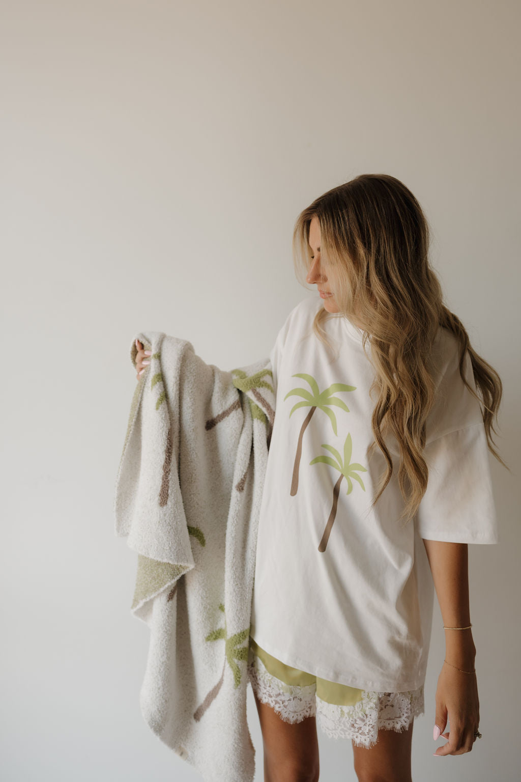 A woman with long, wavy hair holds a palm tree-patterned blanket. She wears the forever french baby Adult Cotton Tee | Palm—a white 100% cotton t-shirt featuring two palm trees—paired with lace-trimmed shorts. The background is plain and neutral.