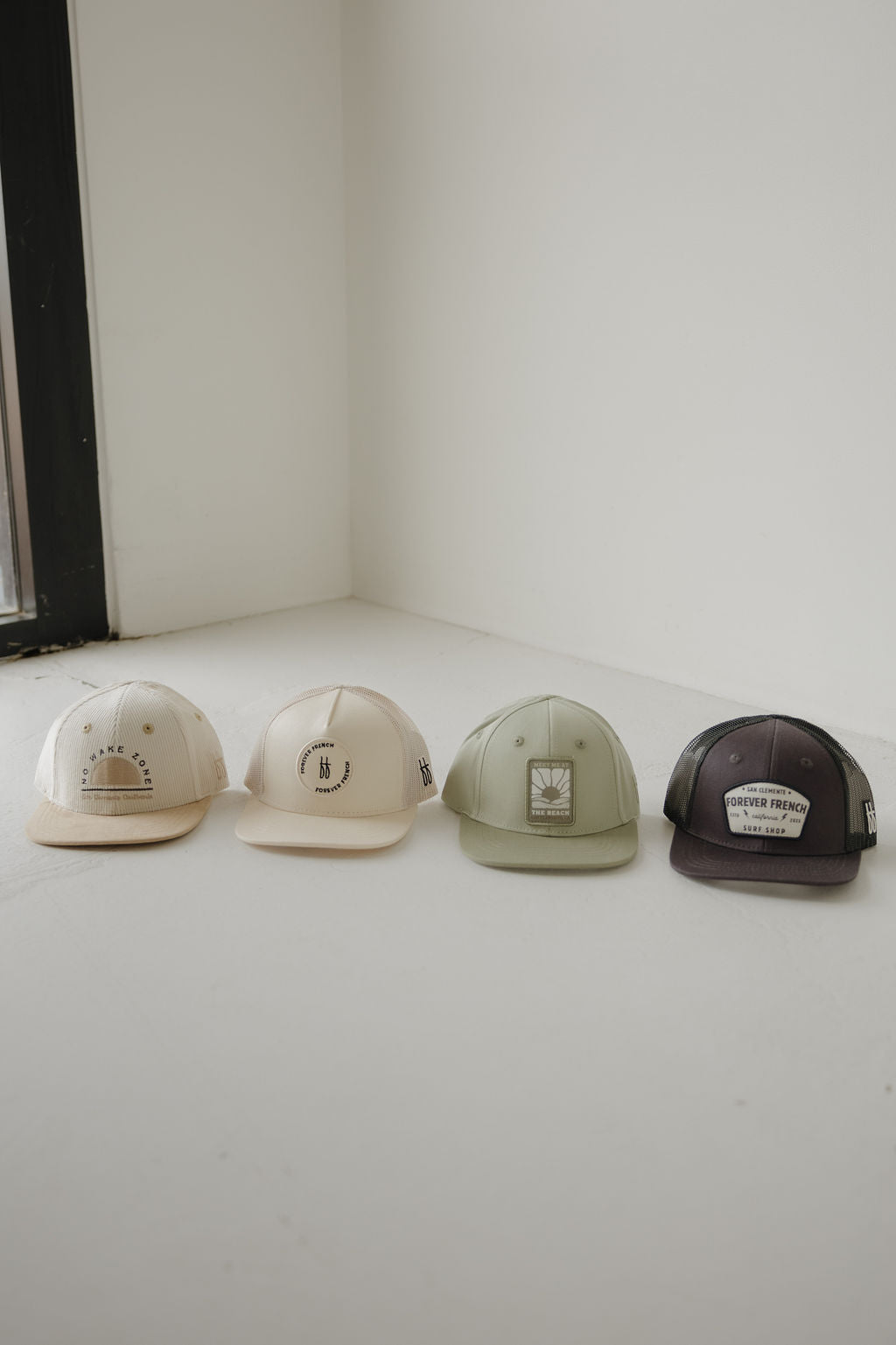 Four forever french baby Snapback | Meet Me at the Beach caps in beige, cream, light green, and black are lined up on a white floor near a window, each featuring its own unique patch or logo on the front.