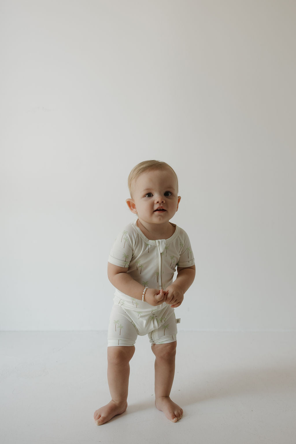 A toddler with short blonde hair stands barefoot on a white floor, wearing the forever french baby Shortie Bamboo Zip in the Palm print—a light-colored, hypoallergenic romper. The background is plain white, and the child gazes slightly upward with a neutral expression.