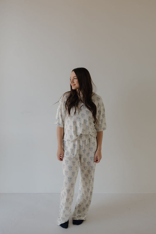 A woman with long dark hair stands against a light wall, wearing the forever french baby Women's Boxy Tee Lounge Set by ff Los Angeles with a subtle floral pattern and dark socks. She looks to her left, appearing relaxed.