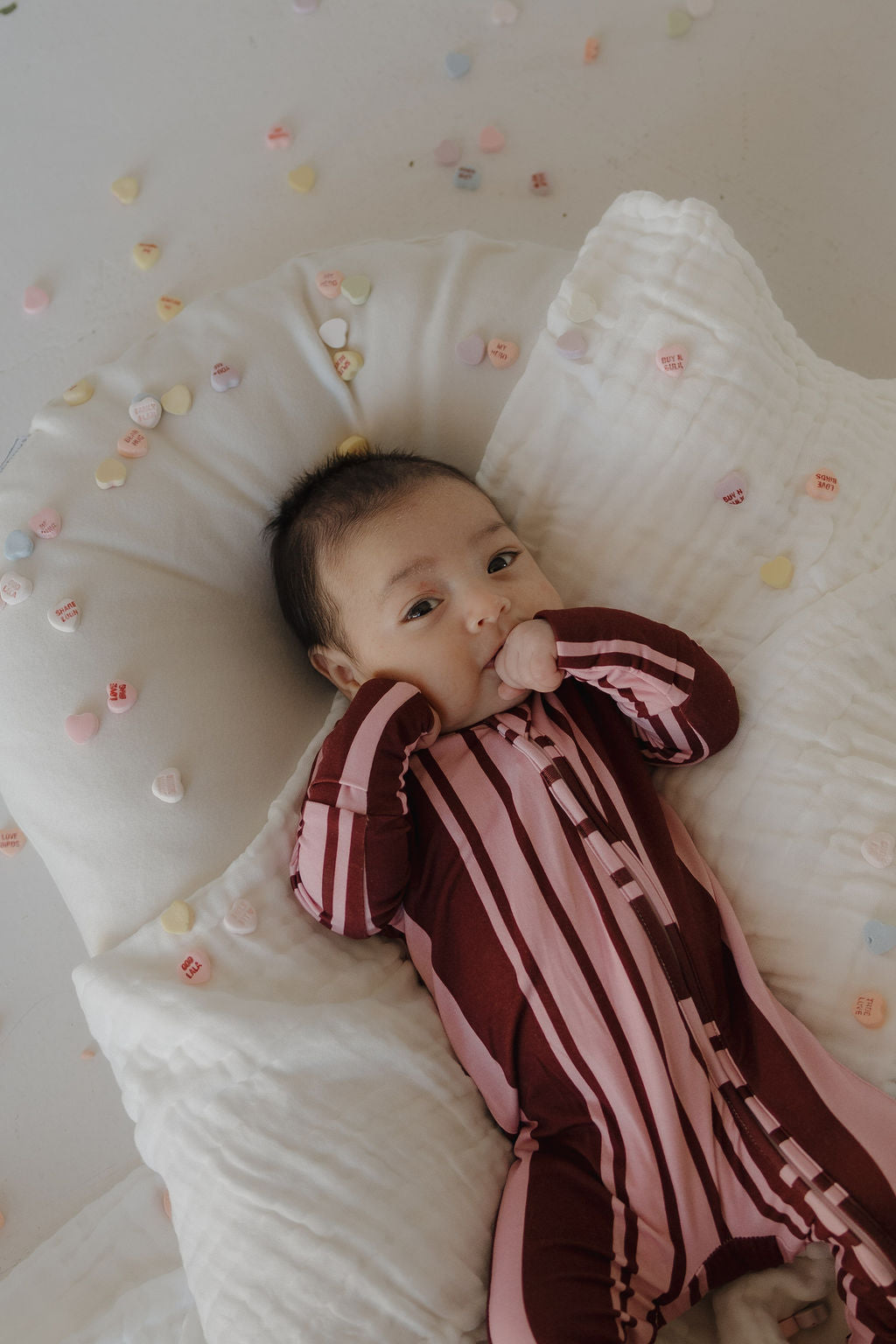 A baby wearing forever french baby's Bamboo Zip Pajamas in Sweetheart Stripes lies on a white pillow, surrounded by pastel candy hearts scattered on a light surface.