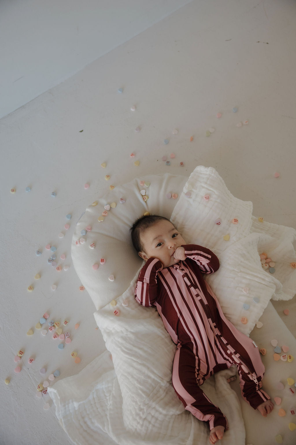 A baby wearing forever french baby’s Bamboo Zip Pajamas in Sweetheart Stripes lies on a white blanket, cozy in the breathable fabric, surrounded by pastel candy hearts as they suck their thumb and look up.