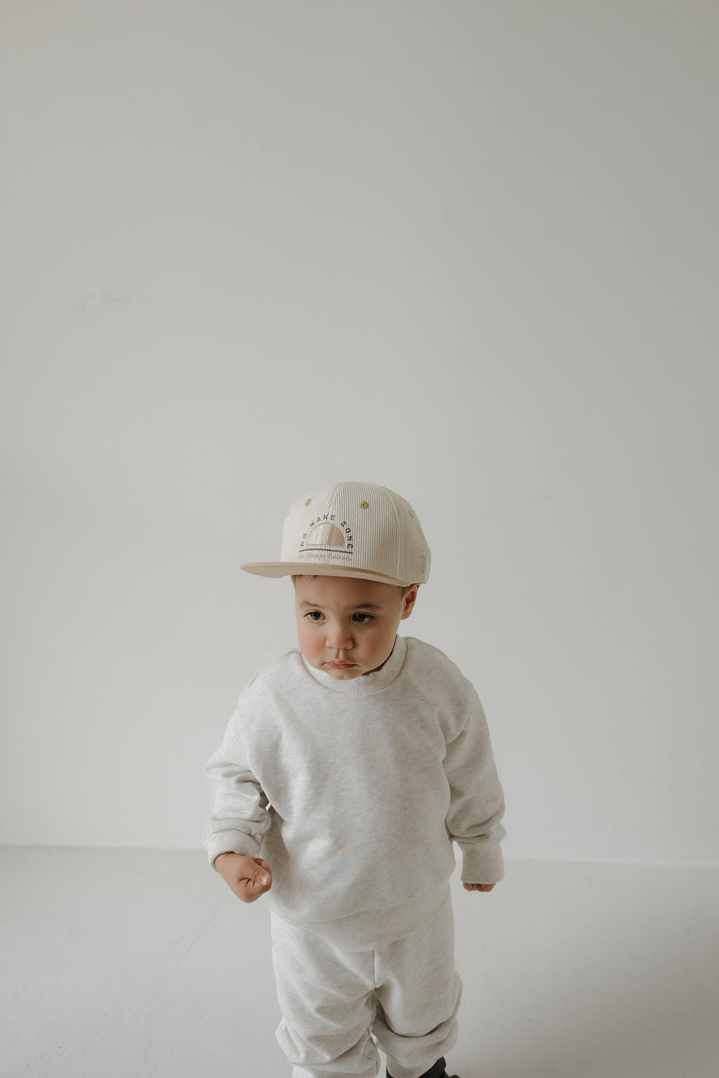 A young child stands against a plain white background, wearing a light sweatshirt, matching pants, and the forever french baby Snapback | No Wake Zone in cream, looking slightly to the side with a neutral expression.