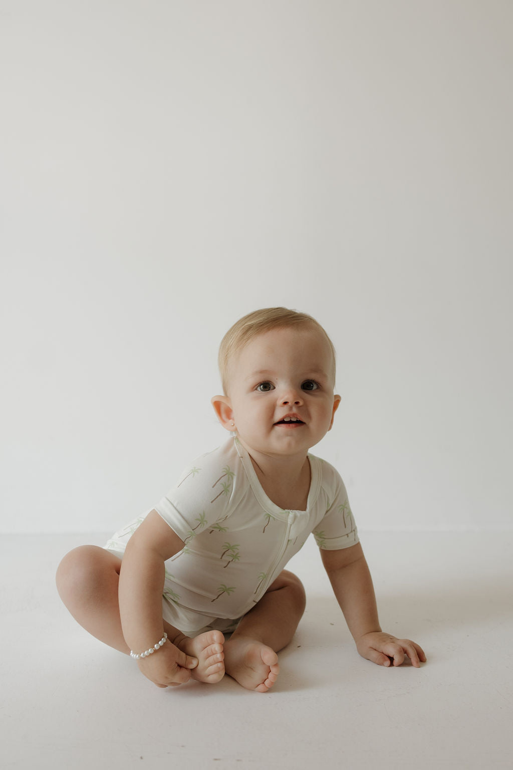 A baby wearing the forever french baby Shortie Bamboo Zip in Palm sits on the floor against a plain white background, looking slightly upward and smiling.