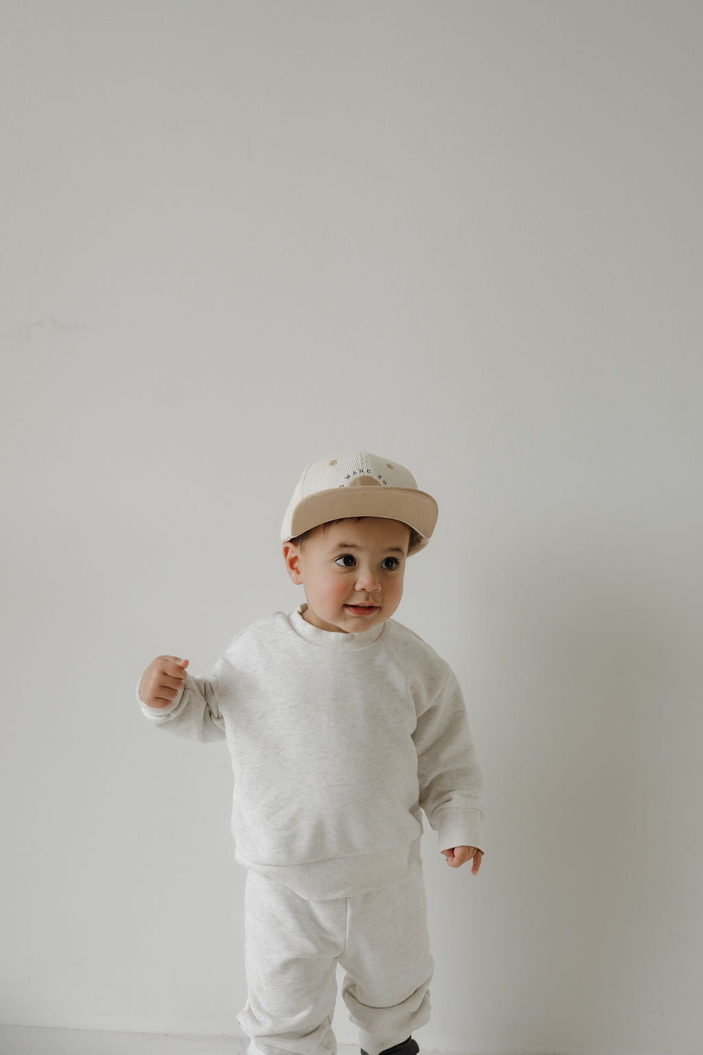 A toddler wears a "No Wake Zone" Snapback from forever french baby, standing against a plain white background and looking to the side with a curious expression.