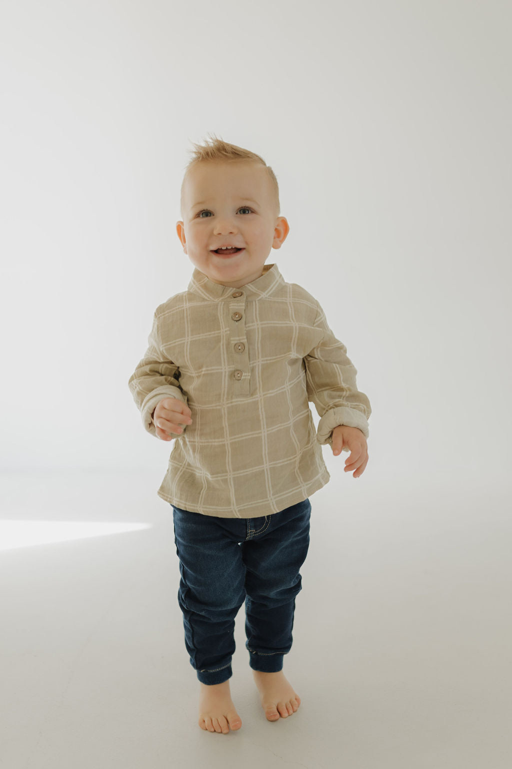 A smiling toddler with short blonde hair stands barefoot on a light floor, wearing forever french baby’s Muslin Button Up | Linen Grid shirt and dark blue jeans, against a plain, light background.