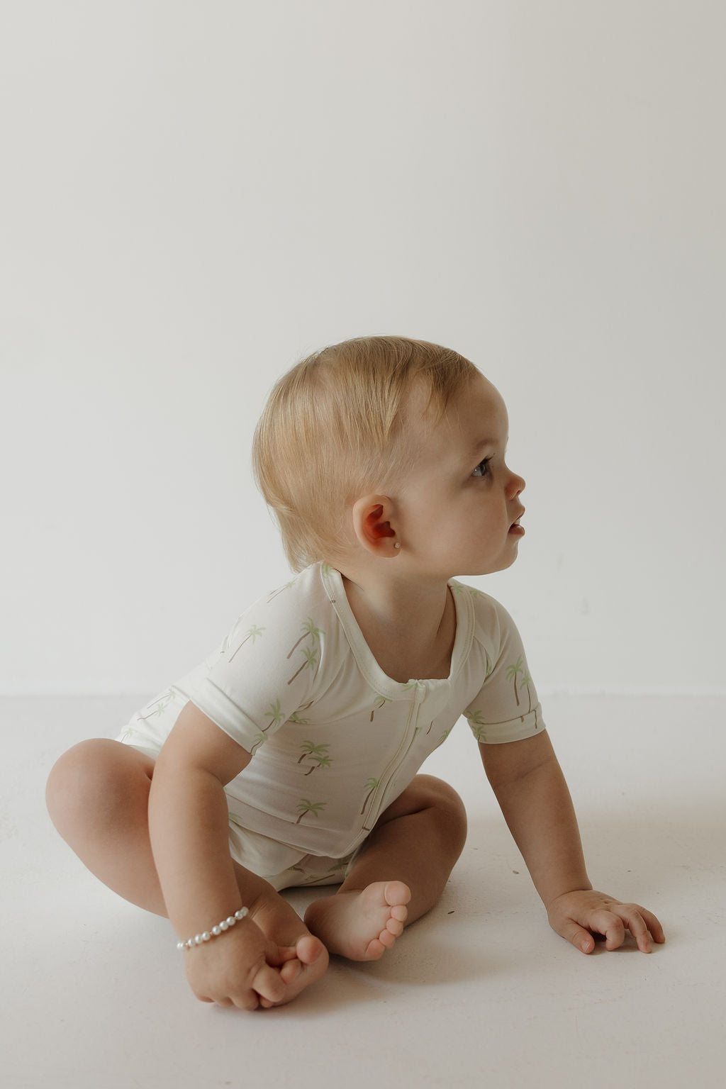 A baby with light hair sits on the floor, looking to the side. The baby is wearing the forever french baby Shortie Bamboo Zip in Palm—a soft, hypoallergenic romper featuring a subtle palm pattern. A small bracelet adorns one wrist, and the gentle scene is set against a plain white background.