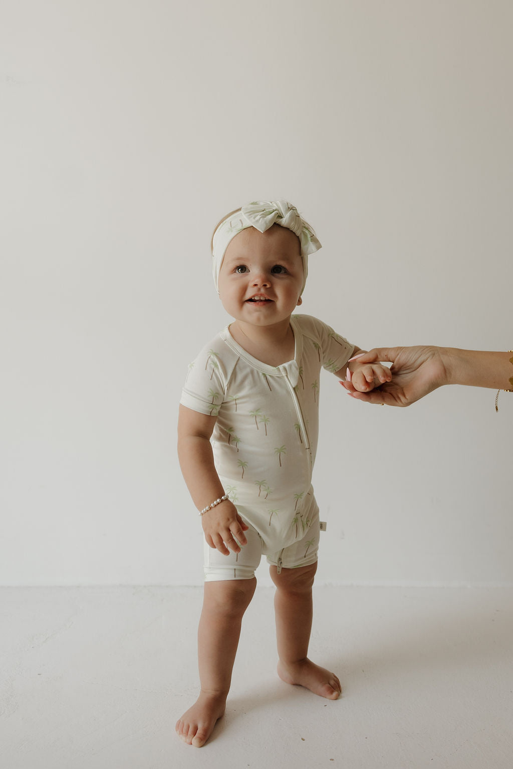 A baby wearing a light-colored onesie and the forever french baby Bamboo Head Wrap | Palm stands barefoot on a white floor, holding an adult’s hand for support and smiling at the camera.