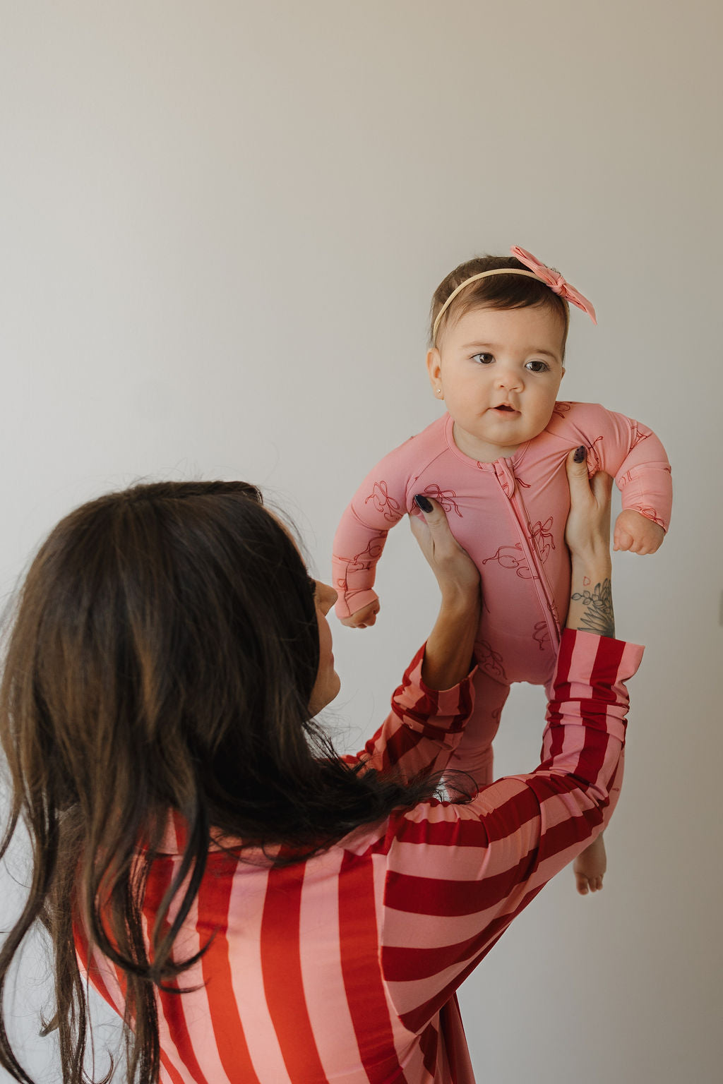 A woman with long dark hair, in a red and pink striped top, lifts a baby in Minty Cherries Bamboo Zip Pajamas | Iconic Cherry and a pink headband. The baby looks forward while the woman faces away against a plain background.
