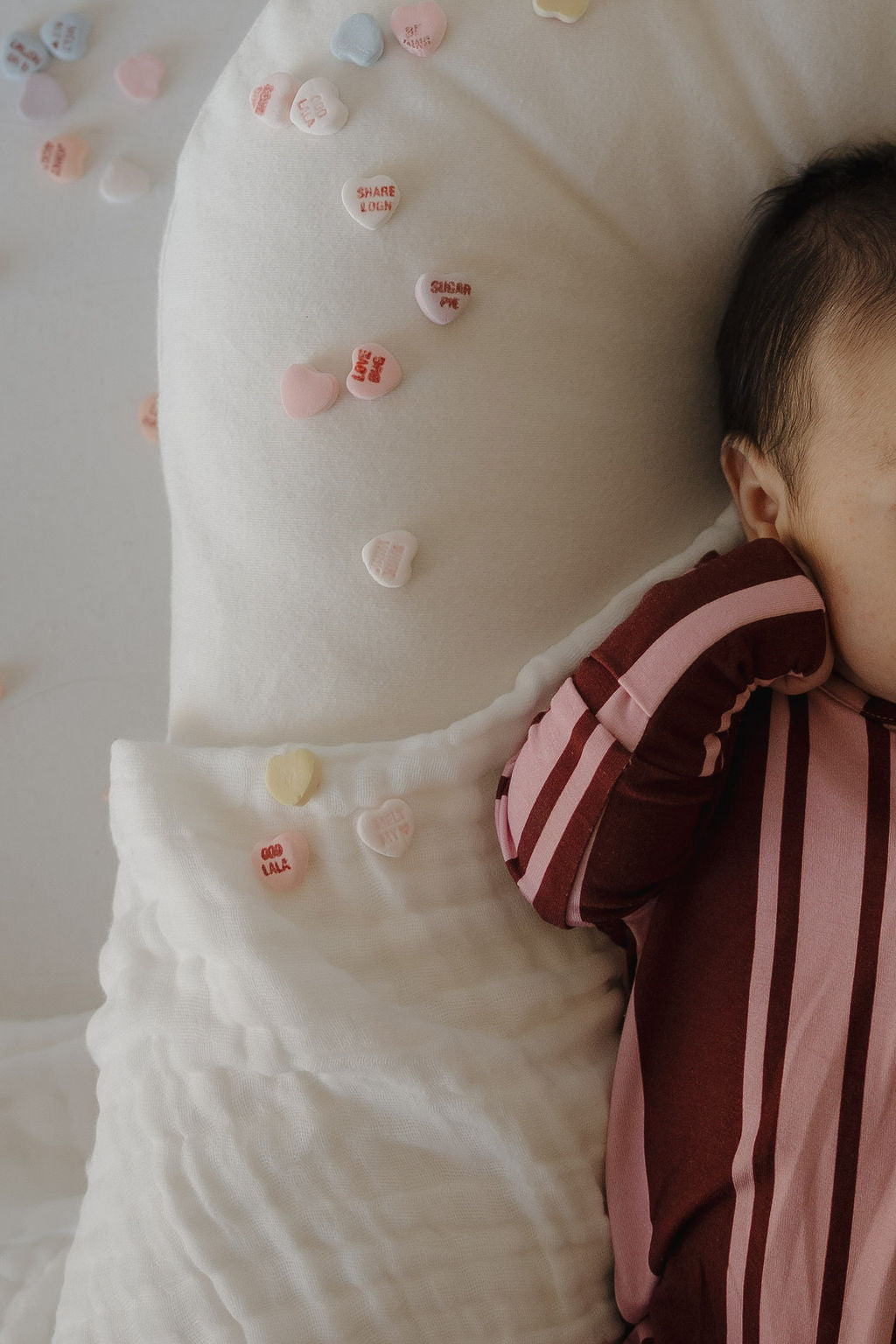 A baby in forever french baby Bamboo Zip Pajamas | Sweetheart Stripes lies on a white blanket, maroon and pink stripes standing out amid pastel candy hearts. Only the right side of the baby's face and body are visible.