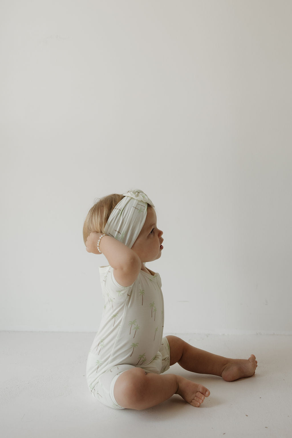 A toddler wearing the Bamboo Head Wrap | Palm by forever french baby sits on the floor against a plain white background, looking to the side with hands touching the stylish head wrap.