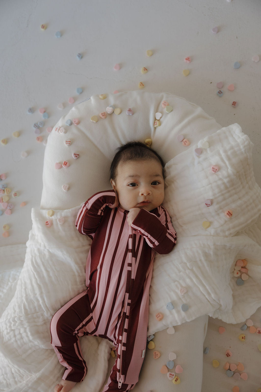 A baby in forever french baby's hypo-allergenic Bamboo Zip Pajamas | Sweetheart Stripes—maroon and pink striped—lies on a white blanket, surrounded by pastel candy hearts.