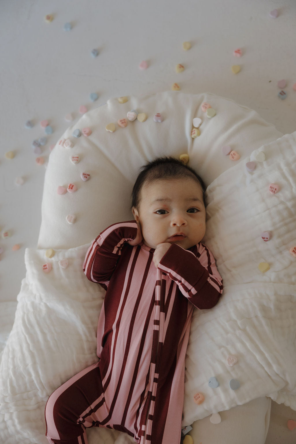 A baby in forever french baby's Bamboo Zip Pajamas | Sweetheart Stripes relaxes on a white cushion, surrounded by pastel candy hearts scattered on the blanket.