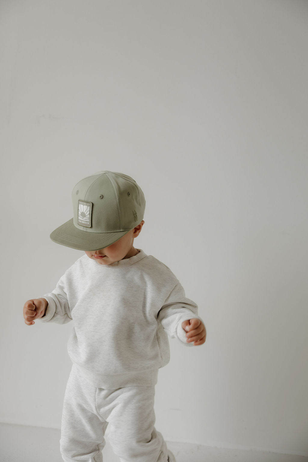 A young child in a forever french baby "Snapback | Meet Me at the Beach" sage green cap and a light gray sweatshirt and pants stands against a plain white background, looking down with arms slightly raised.