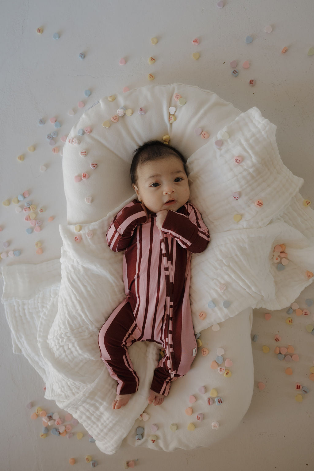 A baby in forever french baby's Bamboo Zip Pajamas | Sweetheart Stripes, featuring maroon and pink stripes, rests on a white cushioned blanket surrounded by pastel candy hearts.
