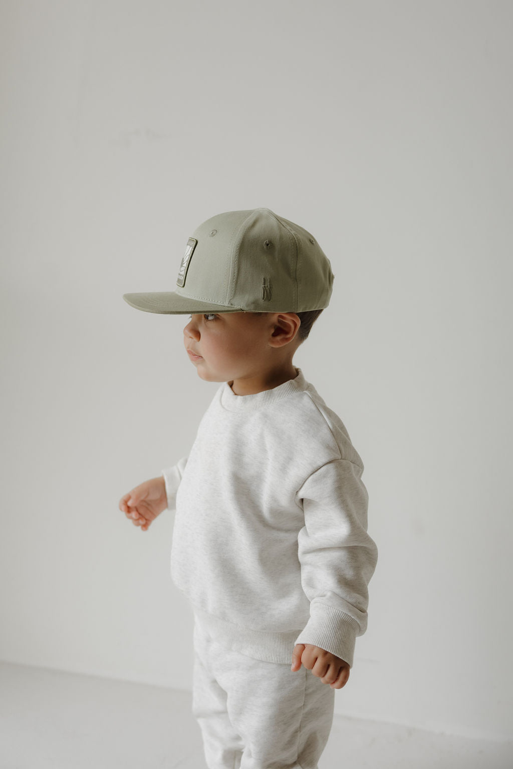 A young child in a light gray sweatshirt and sweatpants stands against a white background, wearing the "Snapback | Meet Me at the Beach" by forever french baby and looking to the left.