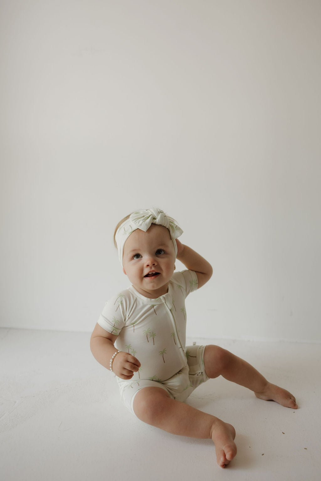 A baby wearing a white onesie and the forever french baby Bamboo Head Wrap in Palm sits on the floor against a plain white background, smiling and touching their head.
