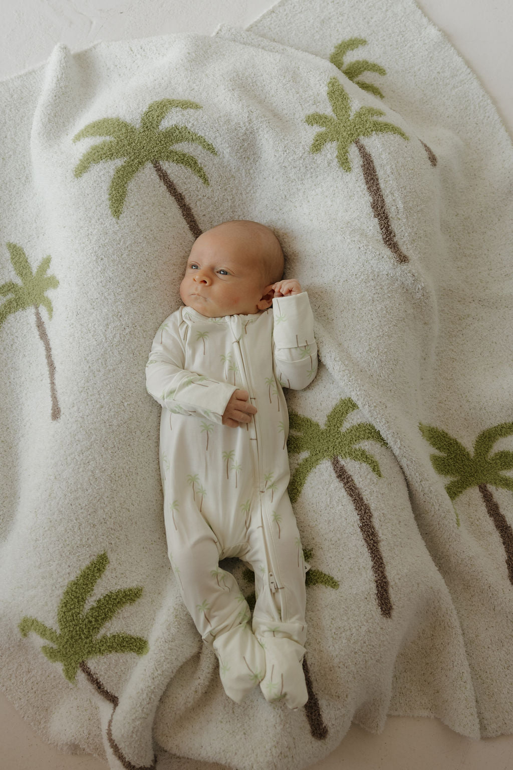 A baby wears forever french baby Bamboo Zip Pajamas | Palm, lying on a cozy white blanket decorated with green and brown palm trees, highlighting the comfort of breathable and hypoallergenic sleepwear.