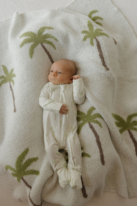 A baby wears forever french baby Bamboo Zip Pajamas | Palm, lying on a cozy white blanket decorated with green and brown palm trees, highlighting the comfort of breathable and hypoallergenic sleepwear.