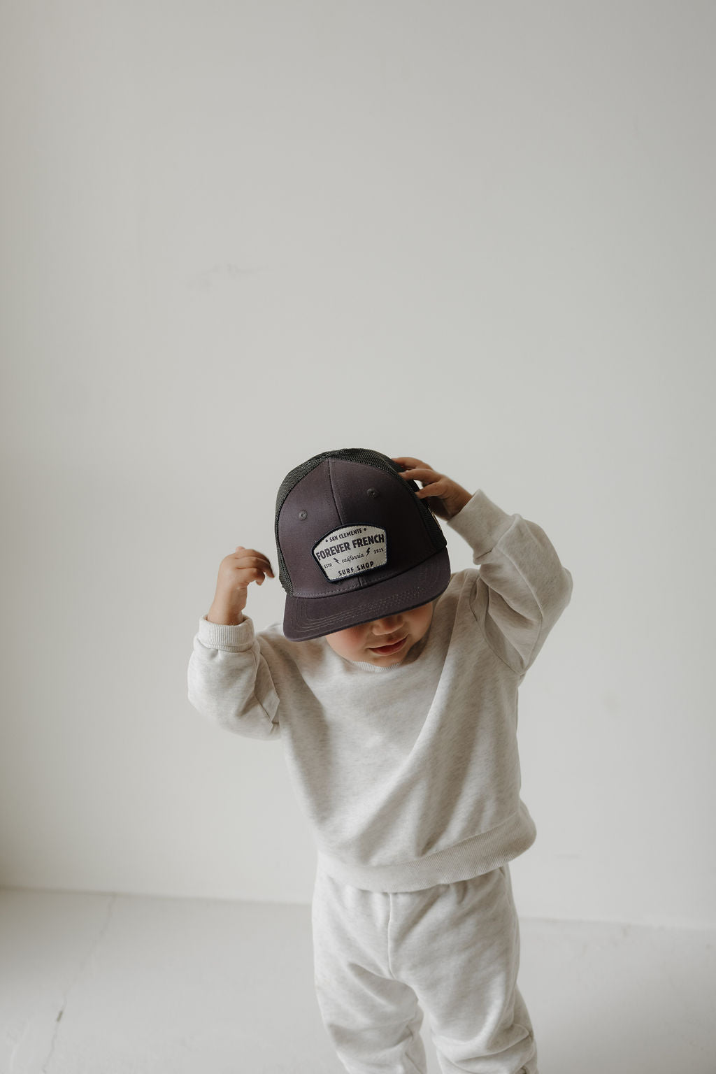 A small child in a light sweatshirt and sweatpants adjusts a large Snapback | ff Surf Shop by forever french baby, standing against a plain white background.