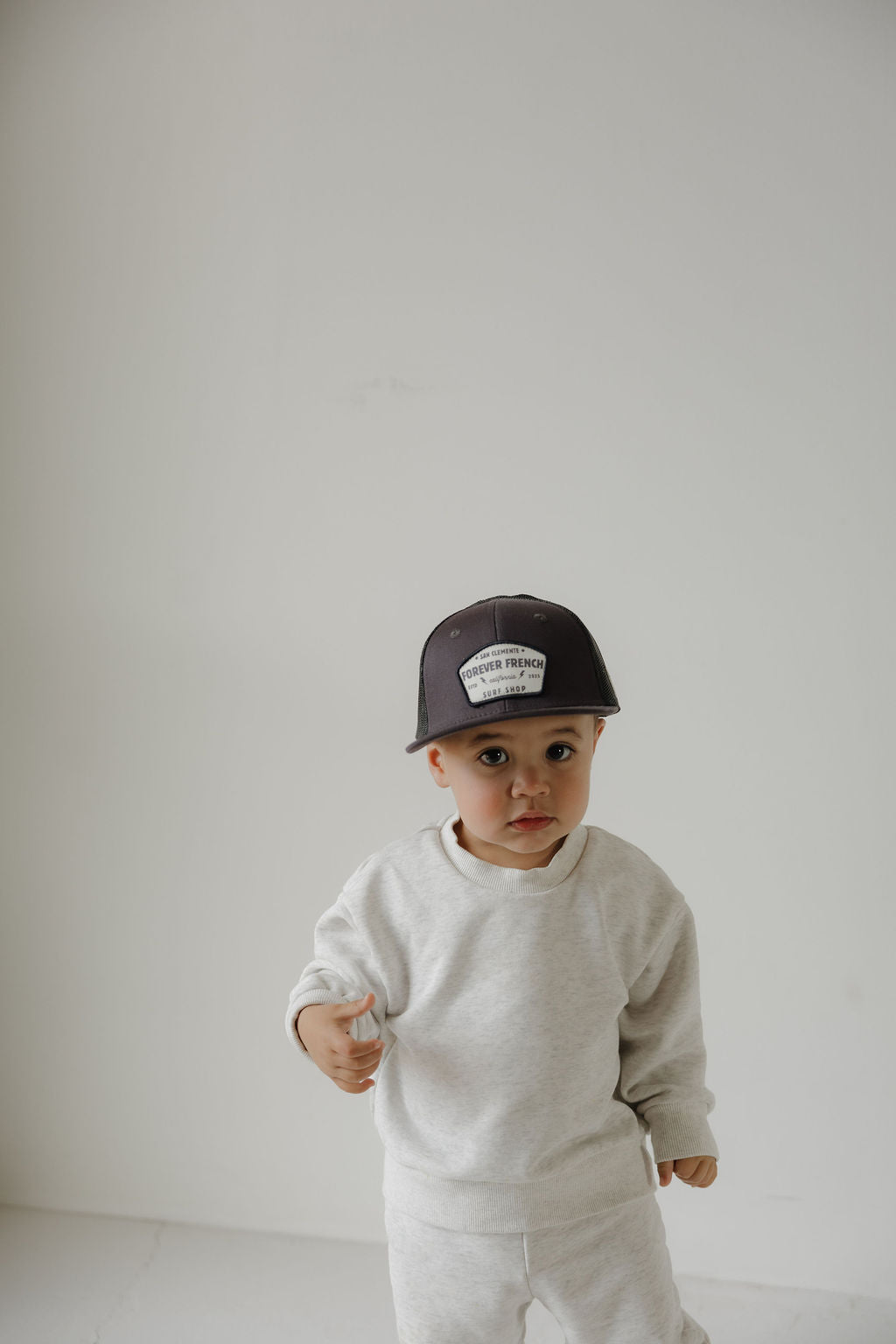 A young child in a light gray sweatshirt and pants stands against a white background, wearing the forever french baby Snapback | ff Surf Shop with a visible front patch, looking at the camera.