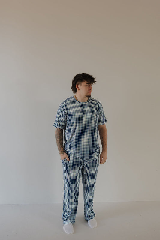 A person stands against a beige wall, wearing the forever french baby Men's Short Sleeve Pajamas in Blue Gingham—hypoallergenic bamboo pajamas for comfort and style—paired with white socks, hands in their pockets.