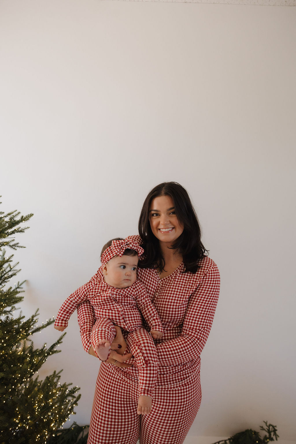 A woman holding a baby, both wearing forever french baby Women's Bamboo Pajamas in Red Gingham, stand by a Christmas tree with white lights against a plain white wall.
