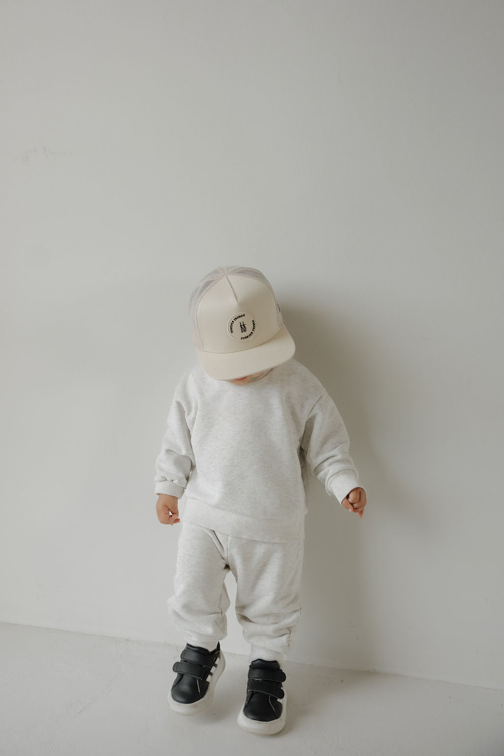 A small child in a light gray sweatshirt, sweatpants, black sneakers, and the Snapback | Sand from forever french baby stands against a white wall, looking down.