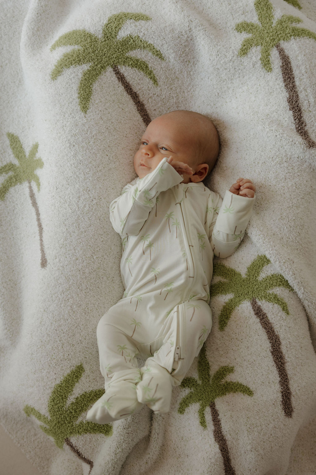 A baby dressed in the forever french baby Bamboo Zip Pajamas | Palm, featuring a white fabric with light green palm designs, lies on a cozy palm tree blanket and gazes to the side with one arm raised—enjoying the ultimate comfort of hypoallergenic baby clothing.
