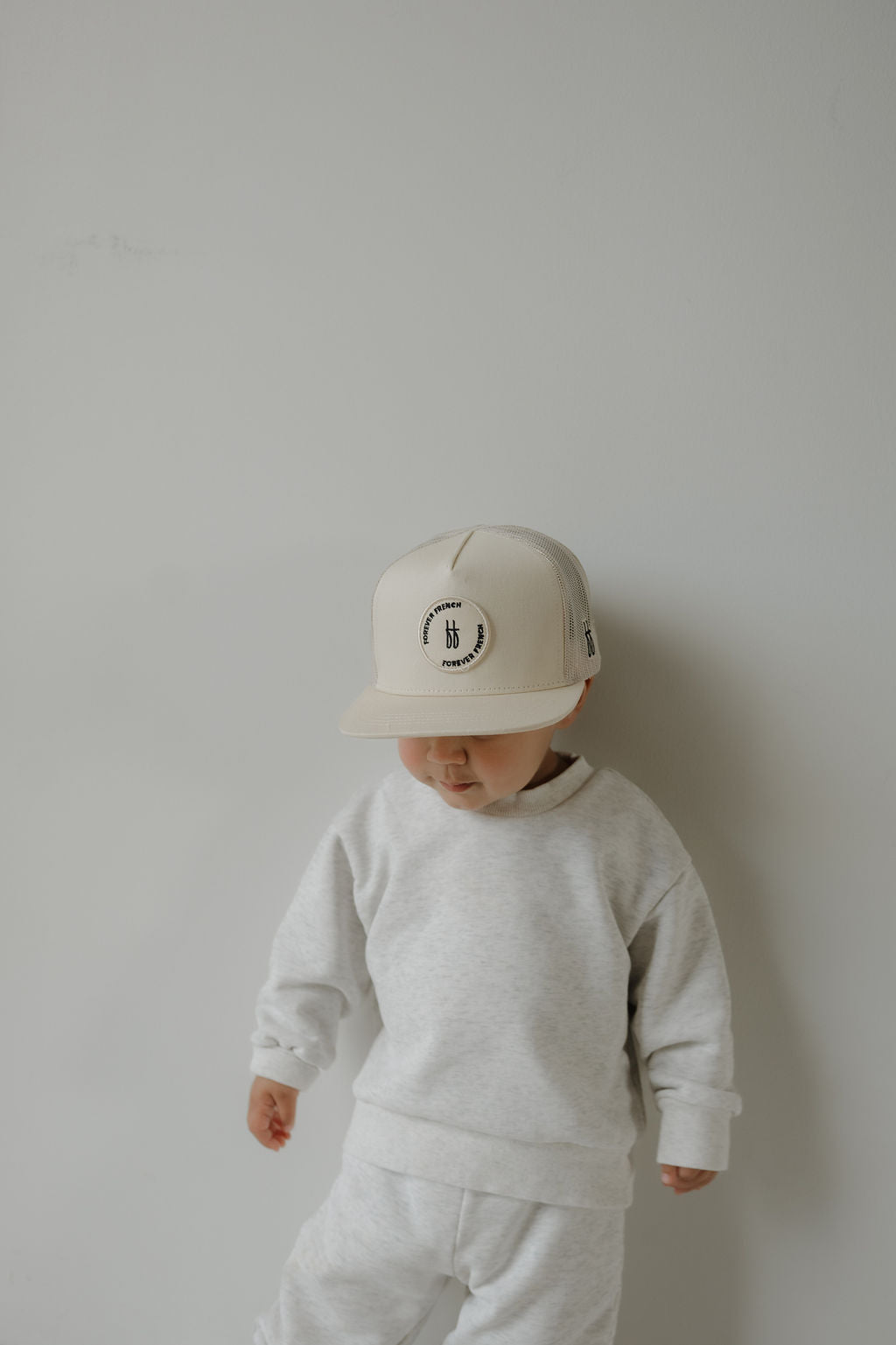 A toddler wearing a Forever French Baby Snapback | Sand and a gray sweatshirt stands against a plain light wall, looking downward.