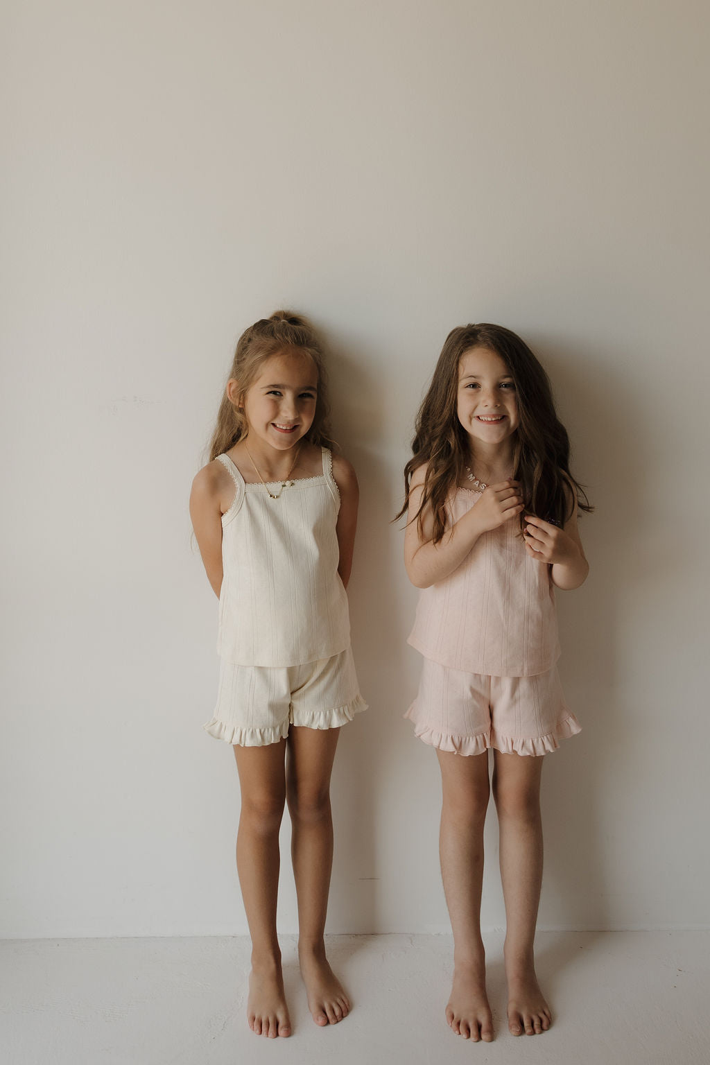 Two young girls with long hair stand barefoot against a plain wall, smiling. One wears a cream-colored pointelle fabric sleeveless top and shorts, while the other is dressed in the forever french baby Girl’s Ruffle Short Set | Swan—a light pink summer outfit featuring a sleeveless top and ruffled shorts.
