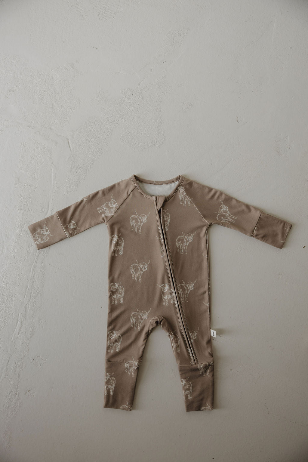 The Bamboo Zip Pajamas | Highland by forever french baby is a brown, long-sleeve romper made from breathable fabric with white animal sketches and a diagonal zipper, displayed flat on a light textured surface.
