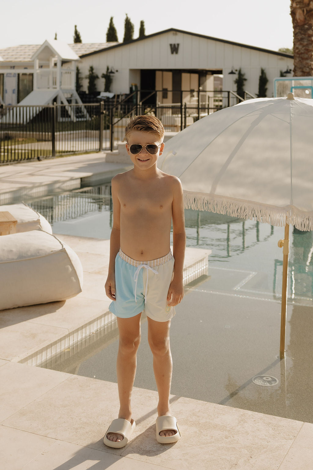 A young boy in lolo webb 2 Child Board Short | Surf & Sand and sunglasses stands by a modern pool under a white umbrella, with lounge chairs and a white building in the sunny background.