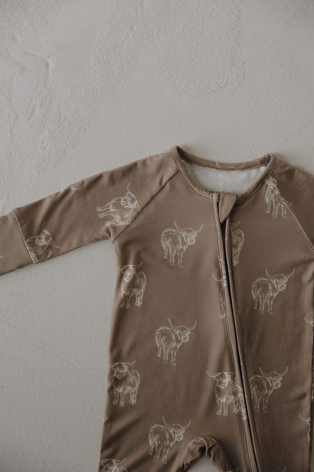 The forever french baby Bamboo Zip Pajamas | Highland is a taupe, hypoallergenic, long-sleeve baby onesie with white highland cow print and a front zipper for easy changing, shown lying flat on a textured surface.