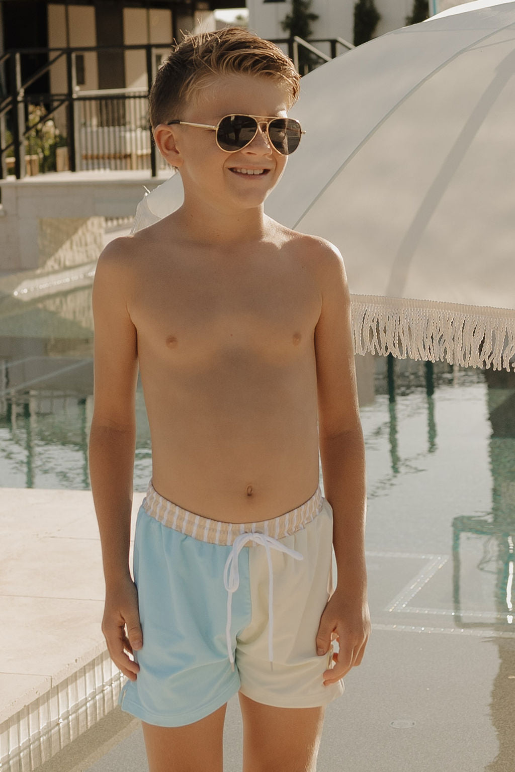 A smiling boy in lolo webb 2's Child Board Short | Surf & Sand and sunglasses stands by a pool with a white umbrella, showcasing a standout from the brand’s limited edition collection.