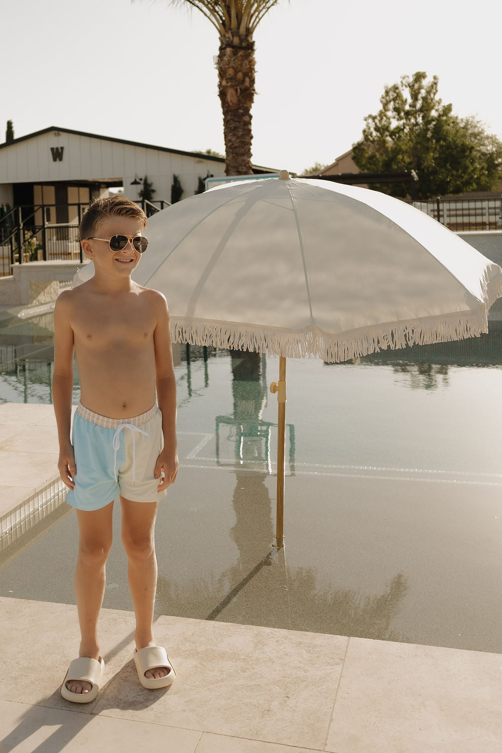 A young boy in lolo webb 2's Child Board Short | Surf & Sand stands poolside under a white umbrella, with sunglasses and slides, as the sun shines on palm trees and modern buildings in the background.