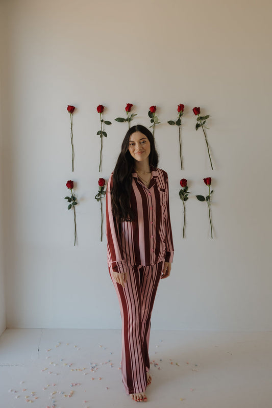 A woman wears the forever french baby Women's Bamboo Button Up Lounge Set in Sweetheart Stripes, standing barefoot in a minimal white room with nine red roses on the wall and petals scattered on the floor.