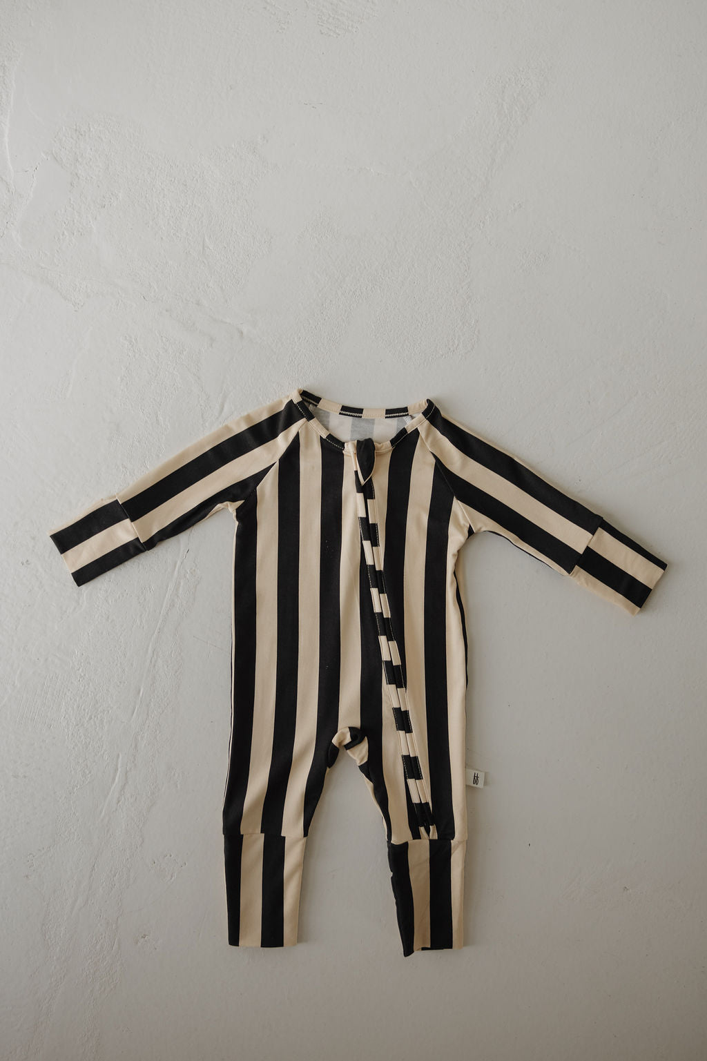 The forever french baby Bamboo Zip Pajamas in Midnight Stripe, featuring black and beige vertical stripes, are laid flat on a textured surface. These breathable pajamas have long sleeves and a front zip for easy changes.