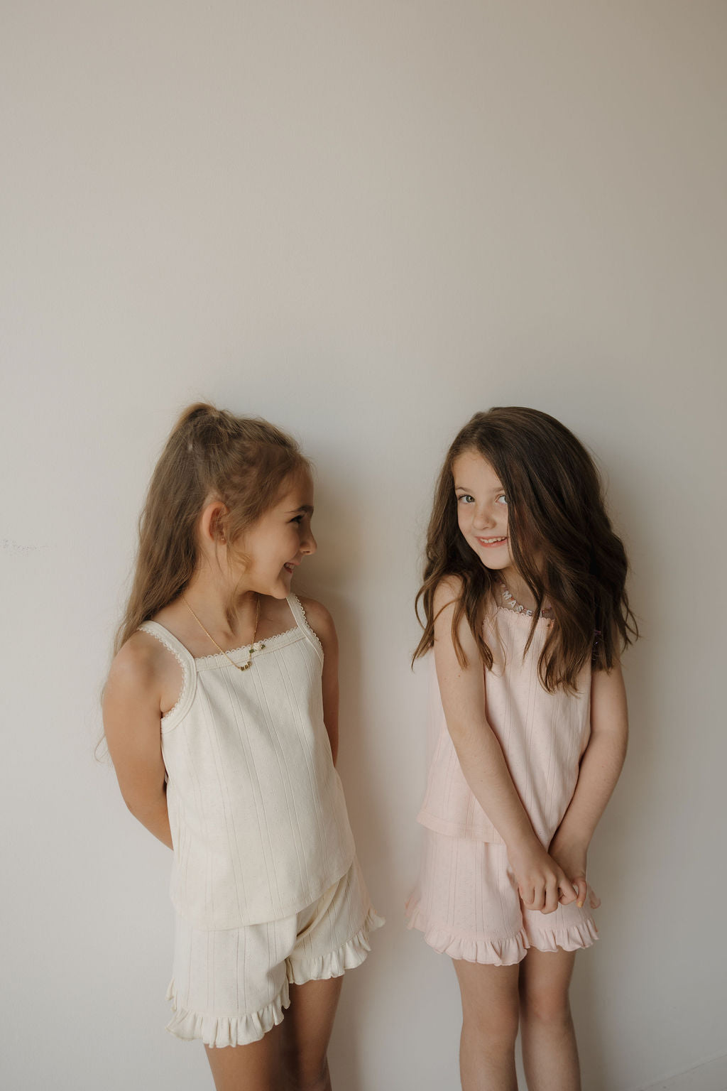 Two young girls with long hair stand against a plain light wall, smiling at each other. One wears a white ruffled outfit made from soft pointelle fabric, while the other wears the Girl’s Ruffle Short Set | Ballerina from forever french baby, crafted from 100% cotton in a light pink hue. Both appear happy and relaxed.