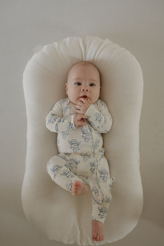 A baby wearing forever french baby's Bamboo Zip Pajamas from ff Los Angeles, featuring soft hypoallergenic fabric with blue animal prints, lies on a cushioned white lounger, gazing up with hands near the mouth and one leg bent.